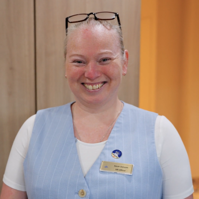 A smiling woman with fair hair pulled back wears glasses on her head, a light blue waistcoat over a white shirt, and a name badge. She stands indoors in front of a wooden background.
