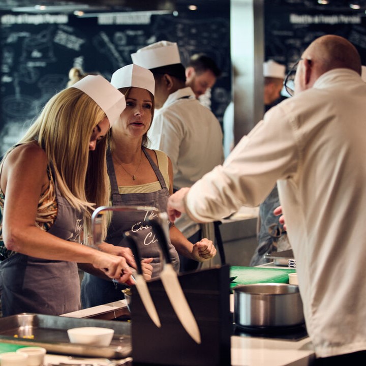 Two women in aprons and chef’s hats listen to a chef’s instructions in a busy cruise ship galley, with cooking utensils and ingredients on the counter. Other crew members work busily in the background.