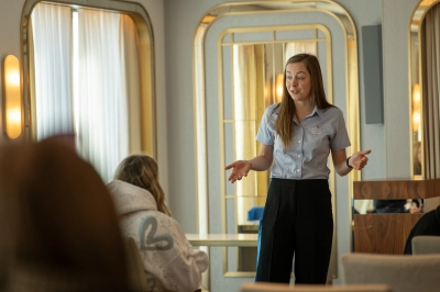 Crew member presenting by a large window inside a lounge area, with curtains and sea view visible outside.