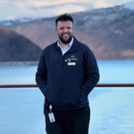 Crew member in a navy jacket standing outdoors on deck, with calm water and mountainous landscape in the distance.