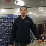 rew member in a storeroom standing in front of shelves stacked with boxed goods and supplies, wearing a dark uniform.