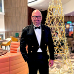 Crew member in a formal dark suit standing indoors beside a tall decorative illuminated structure, with seating and interior décor behind.