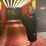 Crew member in a dark uniform standing in a corridor with red walls and lighting, with lift doors visible behind.