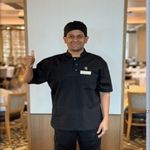 Crew member in a dark uniform standing in a restaurant or lounge setting, with tables, chairs, and warm interior lighting.