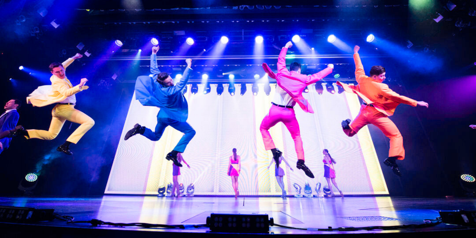 Five performers in colourful suits—yellow, blue, pink, red, and orange—leap energetically on a brightly lit stage, whilst three people stand in the background in front of an illuminated screen.
