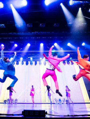 Three dancers in bright blue, pink, and orange suits leap energetically on a brightly lit stage, with spotlights above and other performers in the background.