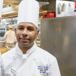 Crew member in a white chef’s jacket and tall toque standing in a bright stainless-steel galley, with cooking equipment and prep surfaces visible behind