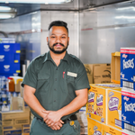 Storekeeper in a green uniform standing in a storage area with stacked boxes and crates arranged on shelves.