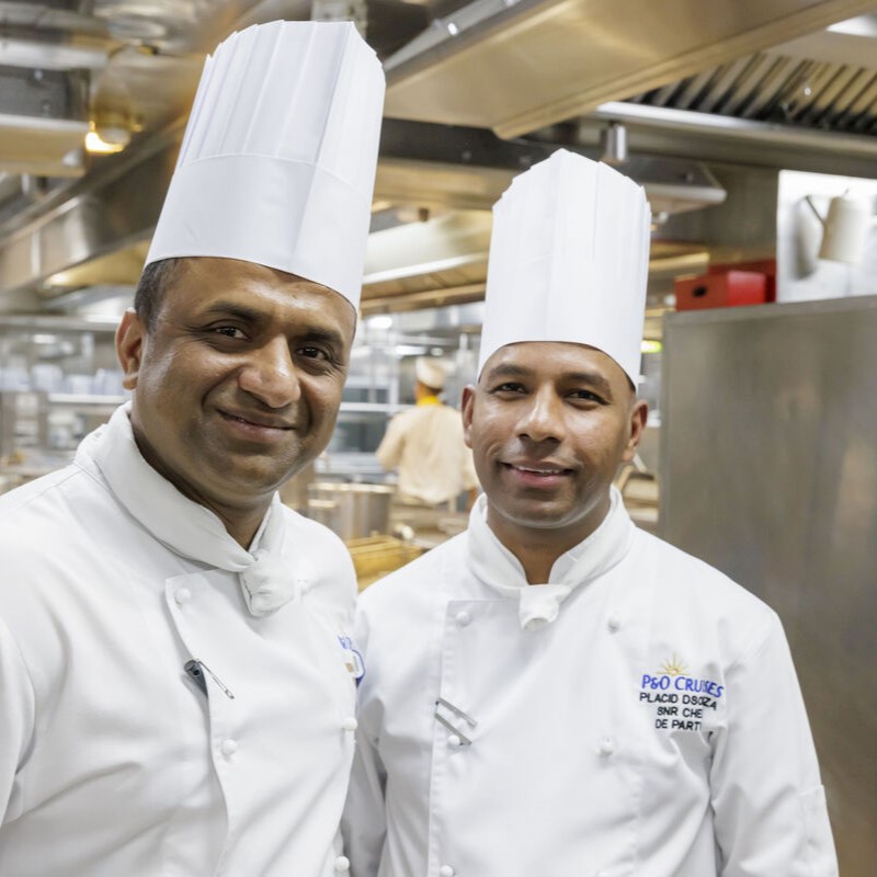 Two kitchen crew members in white chef uniforms and tall hats standing side by side in a well-lit galley with metal counters and equipment.