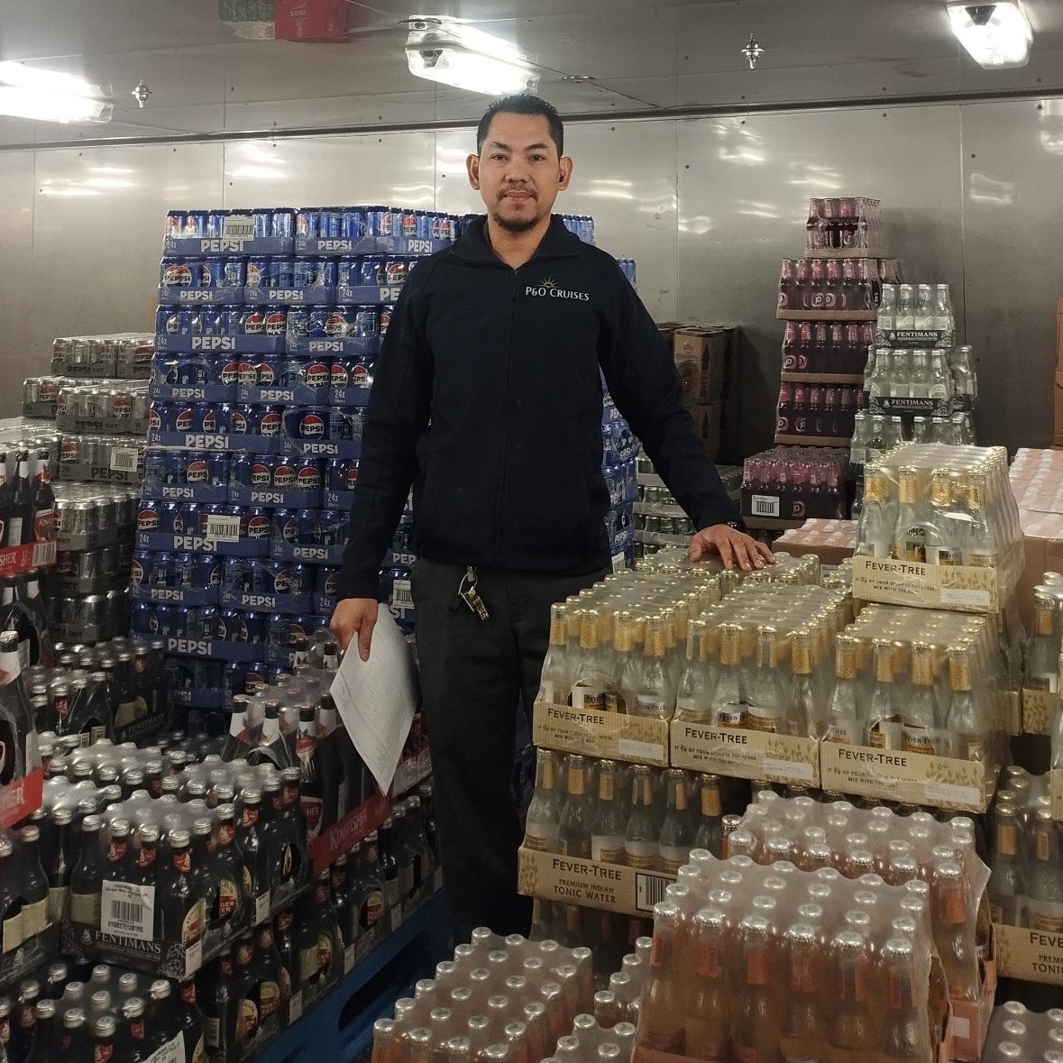 Crew member in dark clothing standing among stacked pallets, bottled goods, and bulk supplies in a large storage area.