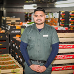 Storekeeper in a green uniform standing in a storeroom aisle, surrounded by shelves stocked with food supplies and packaged goods.