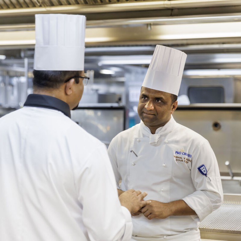 Two chefs in white uniforms facing each other in conversation, surrounded by kitchen workstations and overhead lighting.