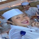 Close-up of a chef in white uniform holding a camera or phone, with other kitchen crew visible in the background of a galley.
