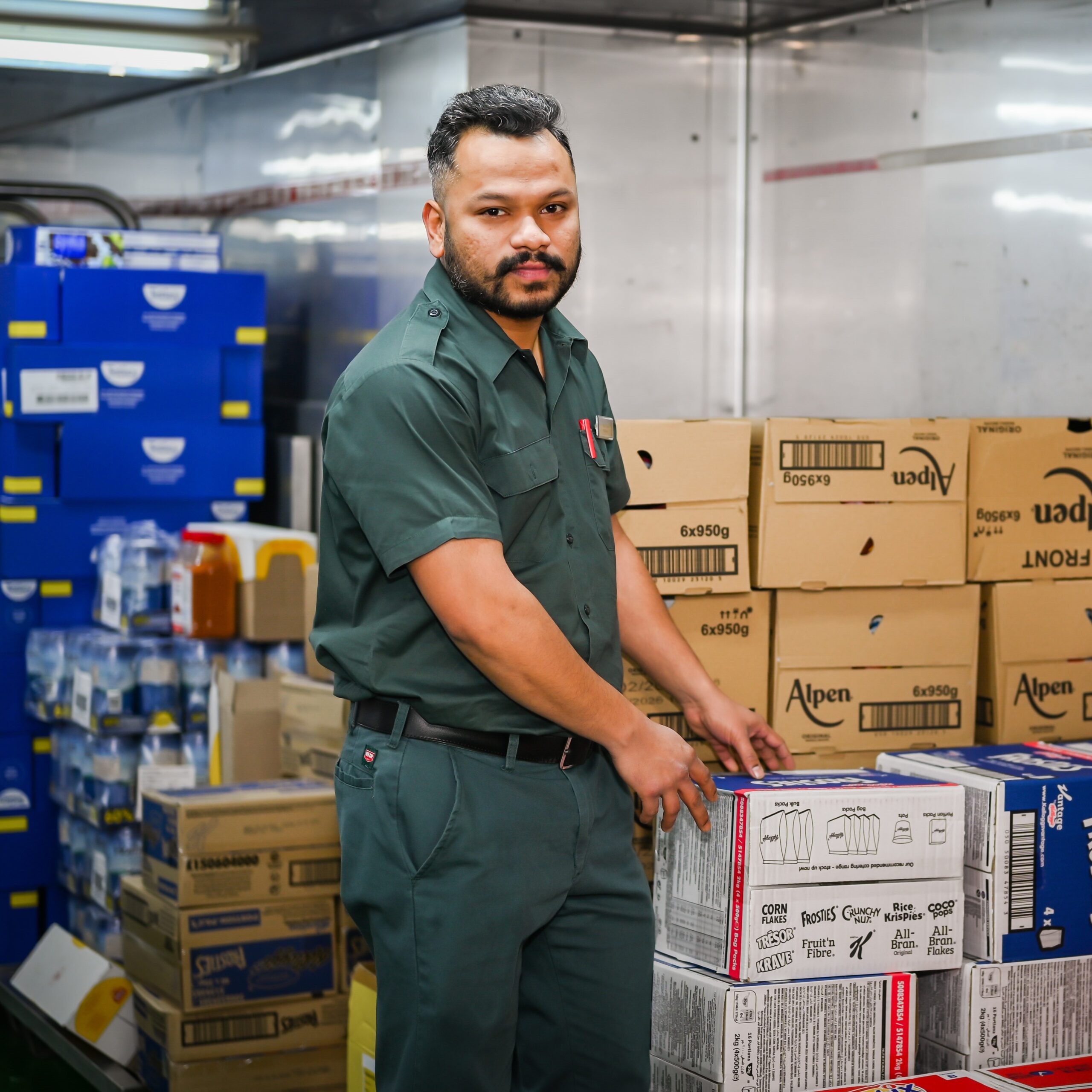 Storekeeper in a green uniform handling or moving boxes in a warehouse-style storeroom with labelled cartons and shelving units.