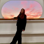 Crew member standing in front of a large porthole with a view of a scenic sunset over the ocean, with soft interior lighting around.