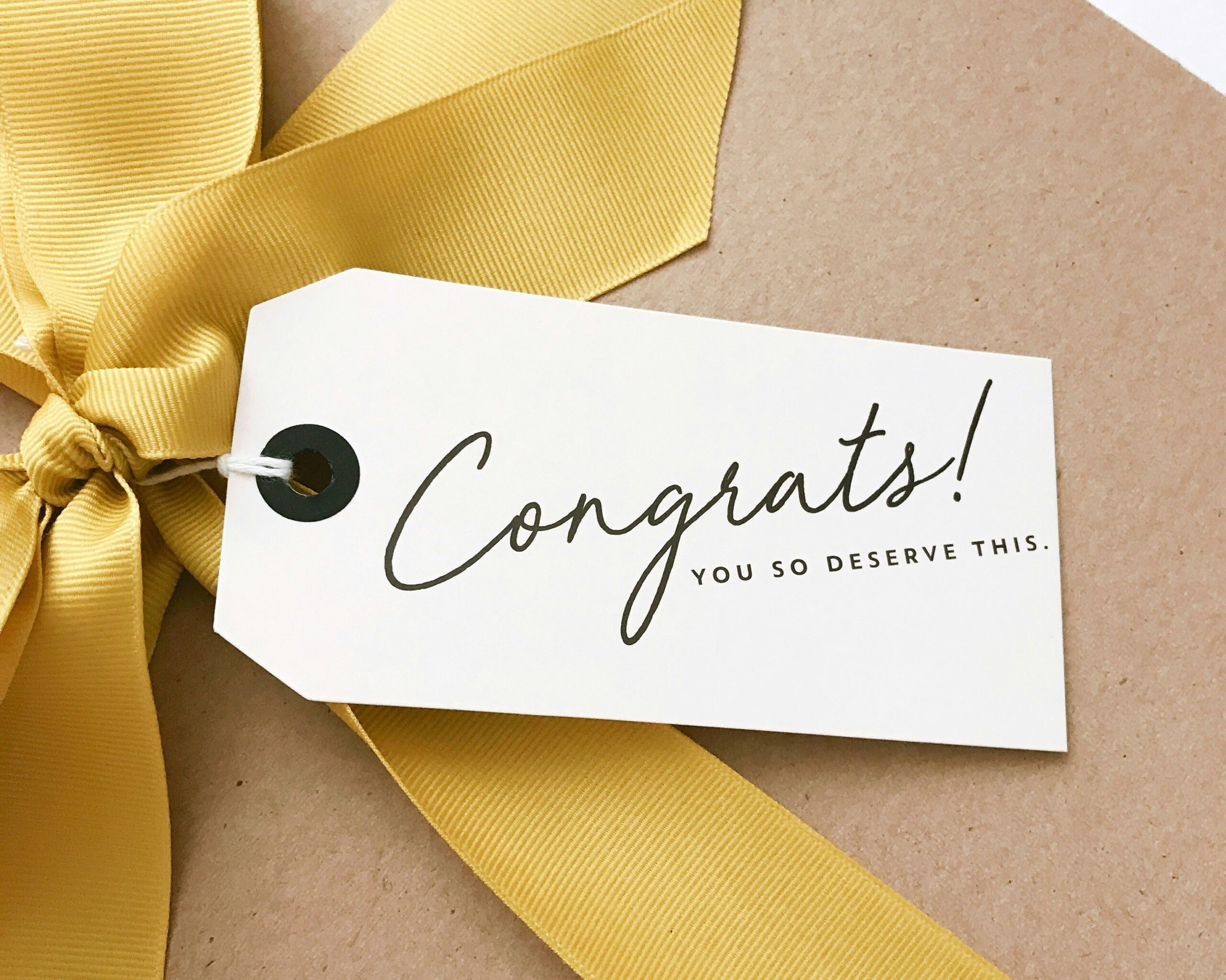A close-up of a present wrapped in brown paper with a large yellow ribbon and a tag that reads, Congratulations! You so deserve this.