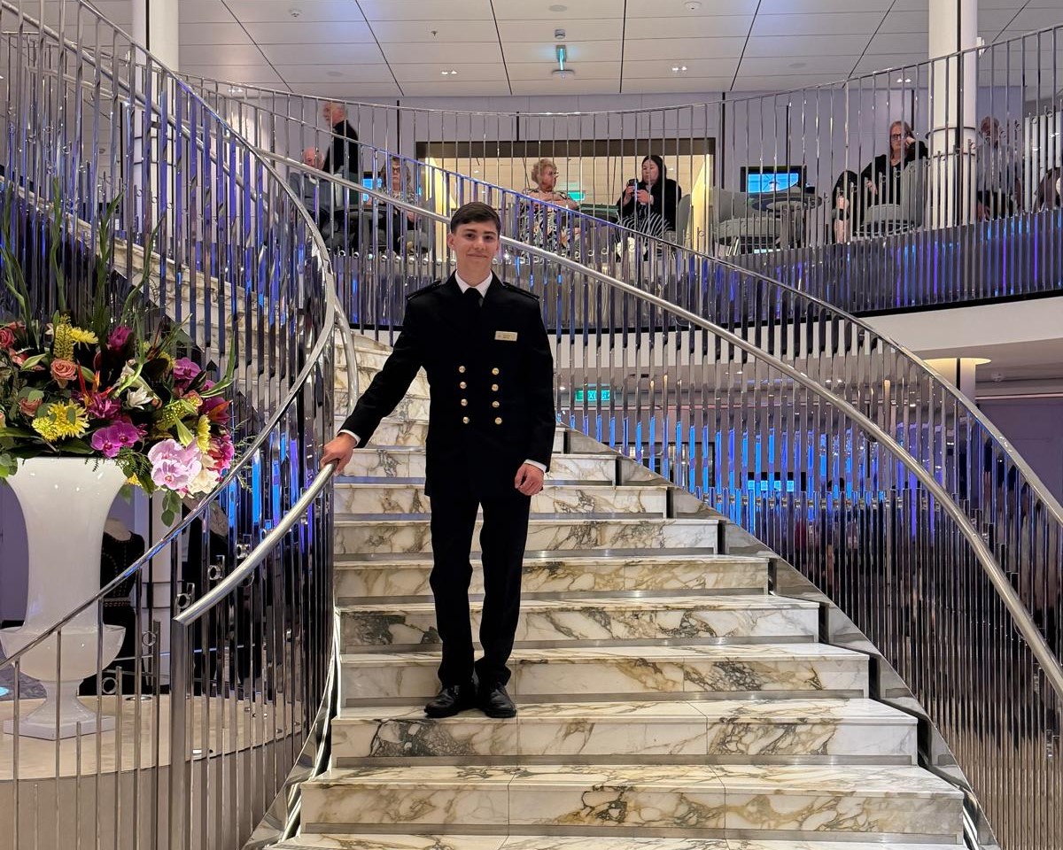 Carnival UK cadet in formal uniform posing on a cruise ship staircase with marble steps and railings, showcasing award‑winning achievement in maritime training and development.