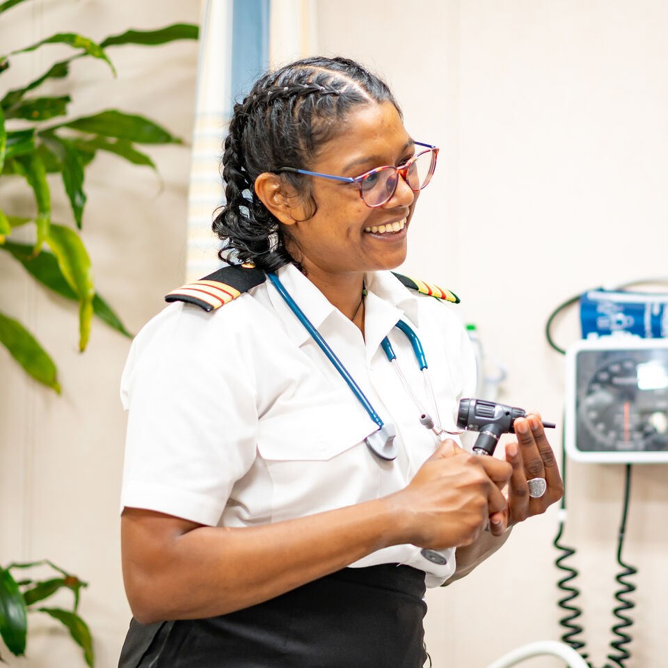 Medical professional in P&O Cruises uniform conducting a health assessment in the ship's medical centre, holding diagnostic equipment and wearing a stethoscope. The image showcases onboard healthcare careers, maritime medical jobs, crew welfare, and medical facilities available on P&O Cruises ships
