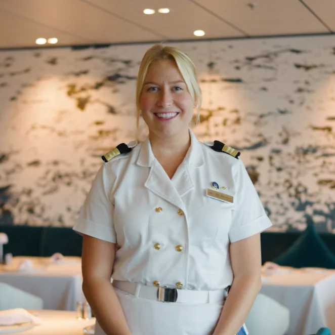 Customer Services Manager onboard a P&O Cruises ship standing in a guest dining venue, wearing a white maritime uniform. The hospitality and guest services professional is positioned in a modern restaurant setting, highlighting customer experience, leadership and onboard operations across cruise ship hotel services at sea.