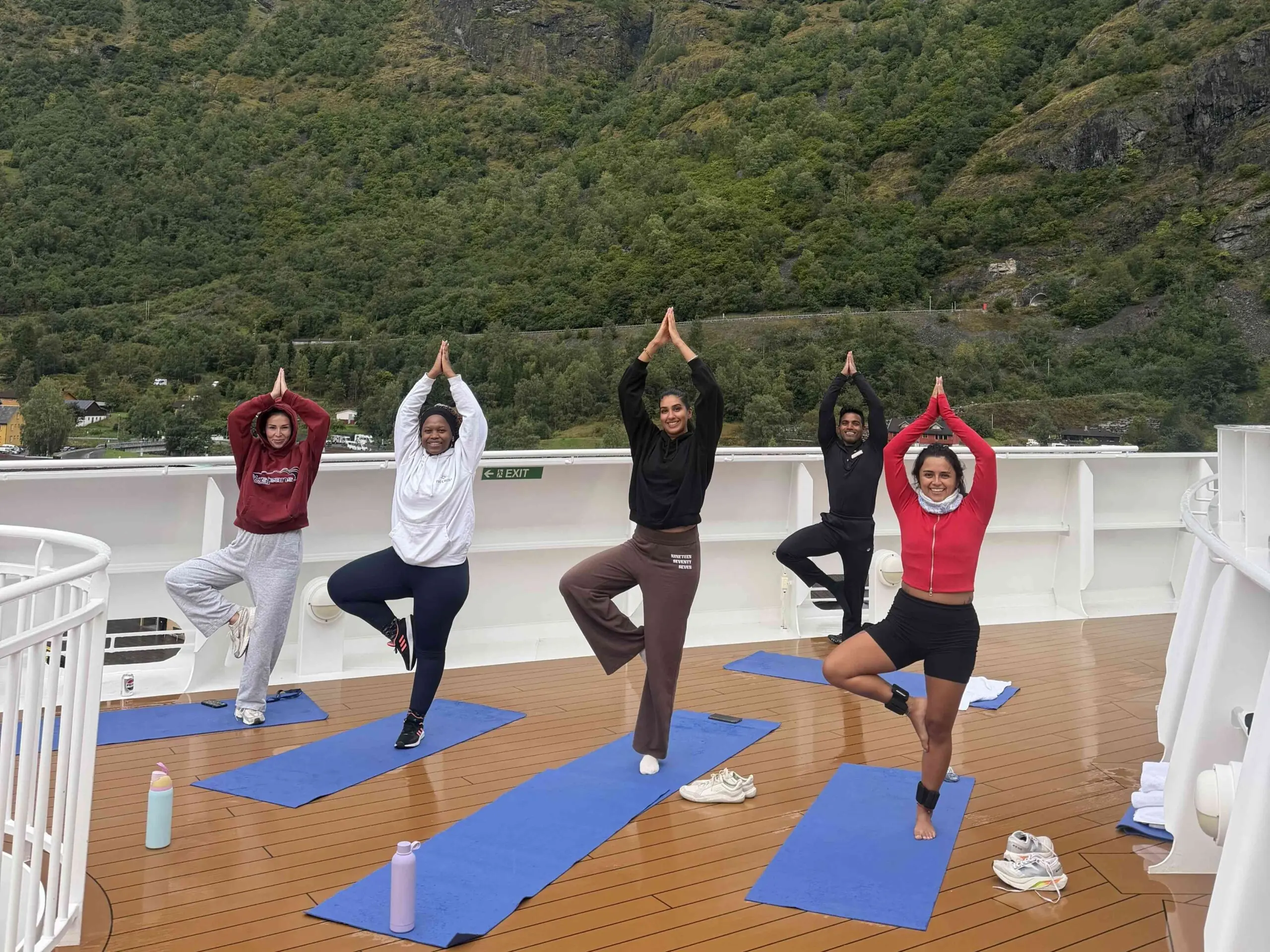Crew members take part in an outdoor yoga session on Arvia's open deck while surrounded by Norway's mountains and fjord scenery.