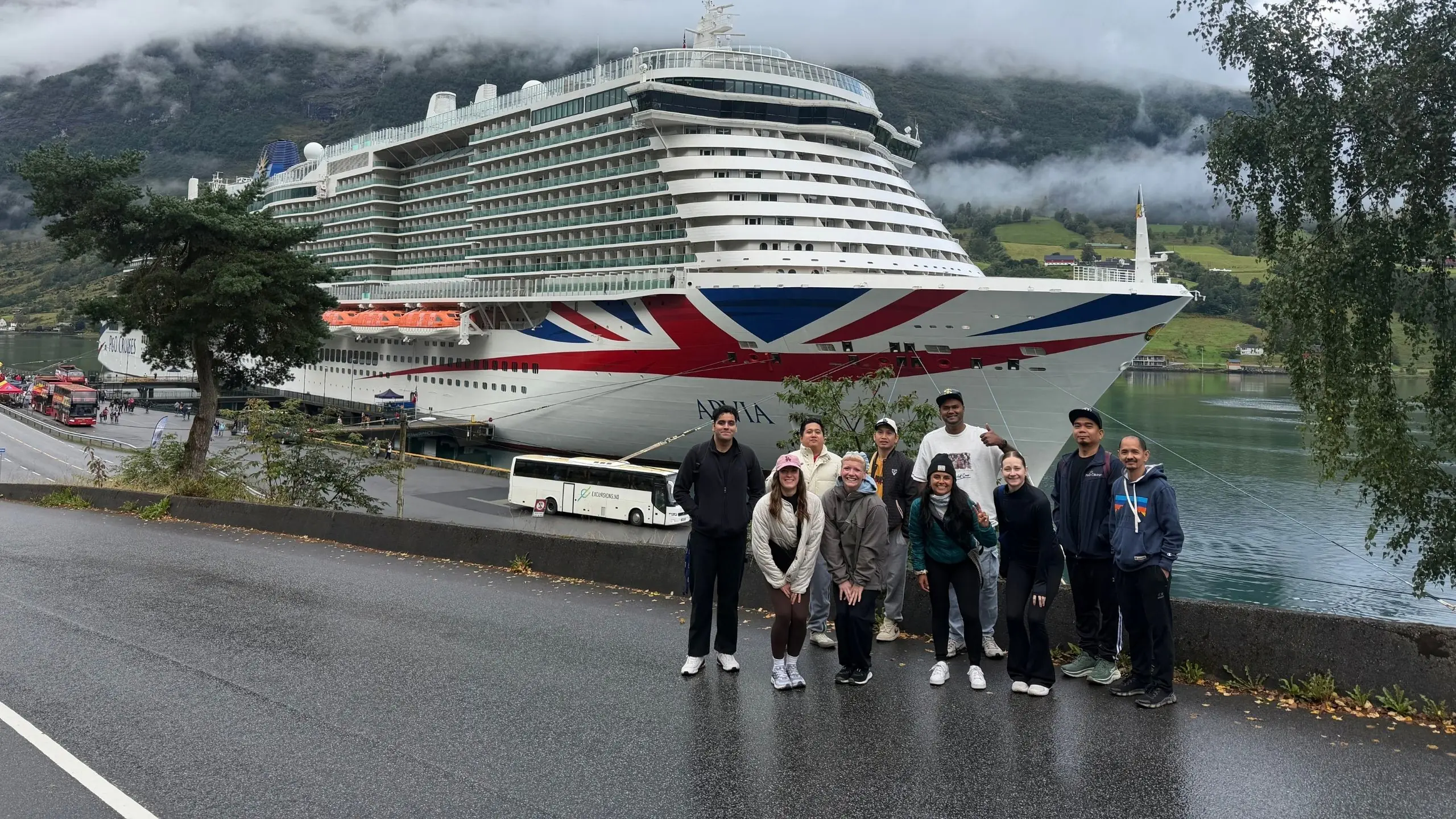 Arvia crew members pose for a group photo during a Norway shore excursion, with Arvia docked beneath mist-covered mountains in the Norwegian fjords.